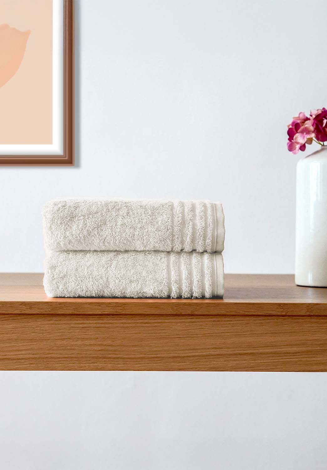 Zwei gefaltete, cremefarbene Handtücher liegen ordentlich auf einer hölzernen Oberfläche. Daneben steht eine weiße Vase mit pinken Blüten, und im Hintergrund hängt ein gerahmtes Kunstbild an einer hellen Wand.