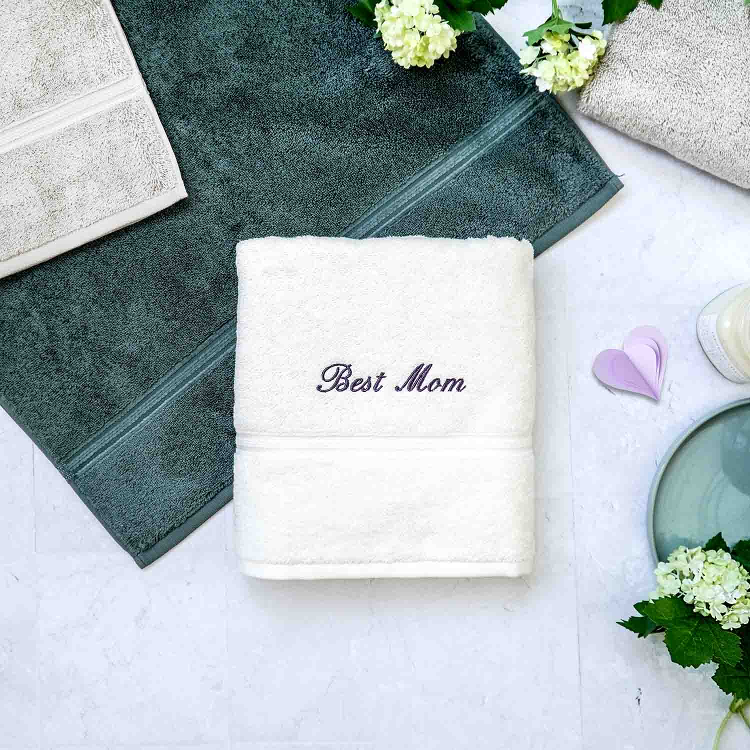 Ein weißes Handtücher mit der Aufschrift ‚Best Mom` auf hellem Hintergrund, daneben Deko.