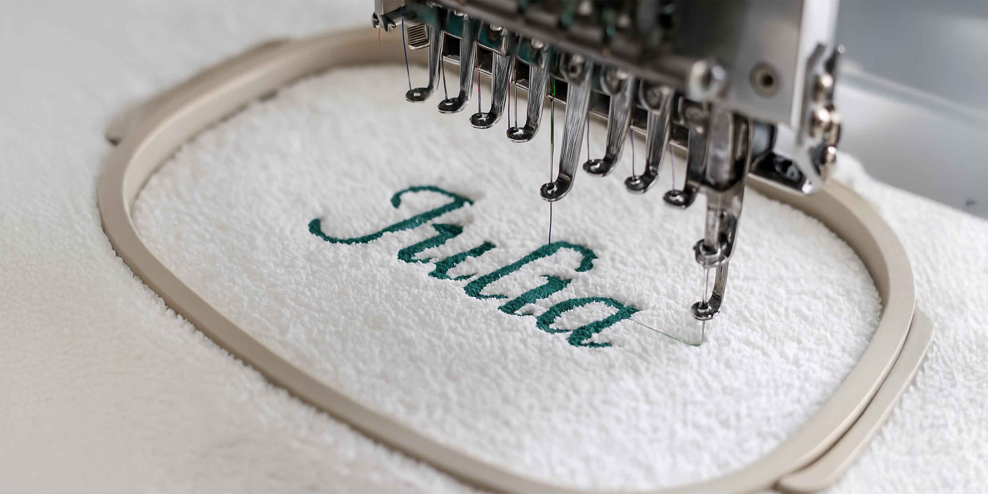 Stickmaschine näht den Namen ‚Julia‘ in grüner Schrift auf weißes Handtuch, eingespannt in einen Stickrahmen.