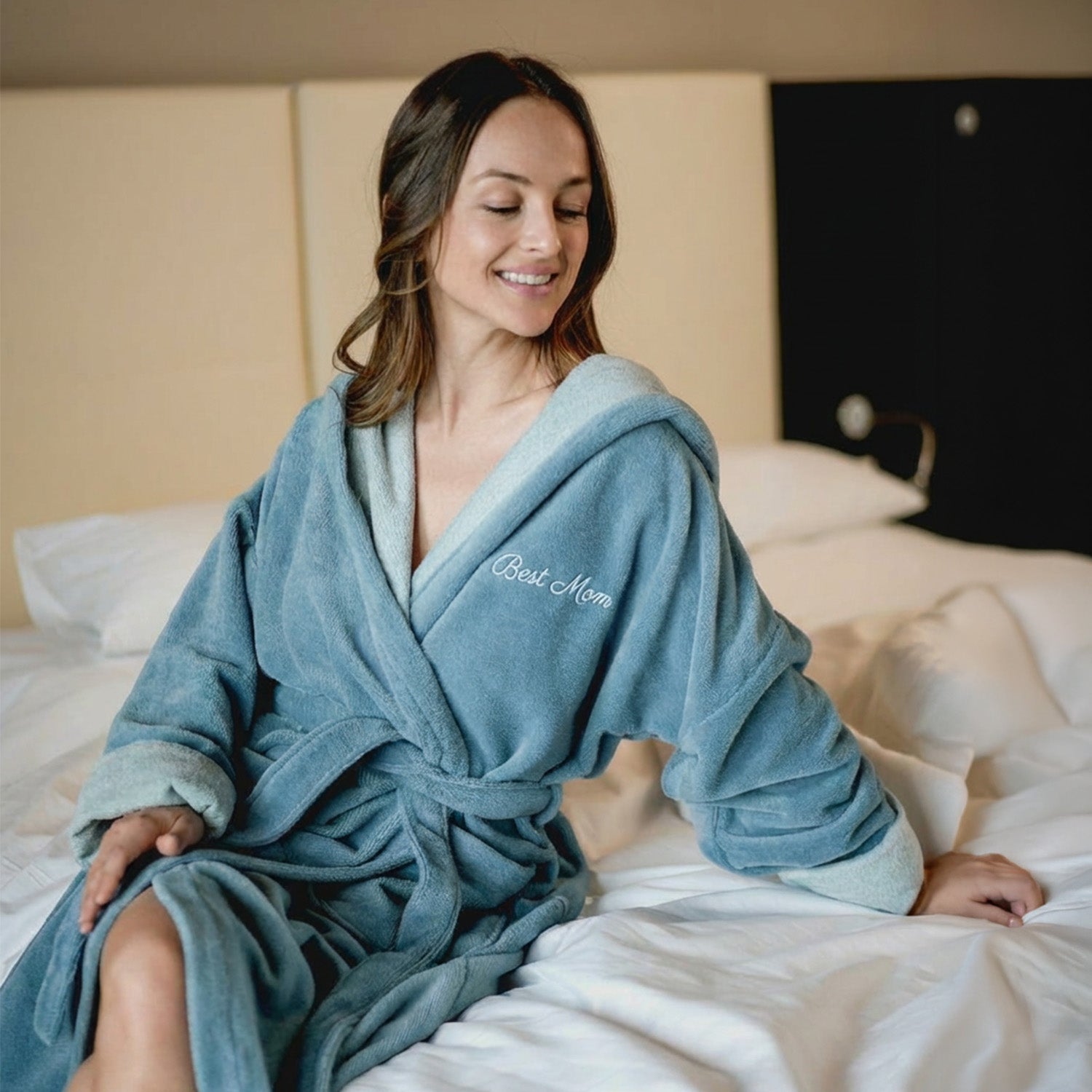 Frau im Bademantel mit Stickerei ‘Best Mom’ sitzt auf dem Bett
