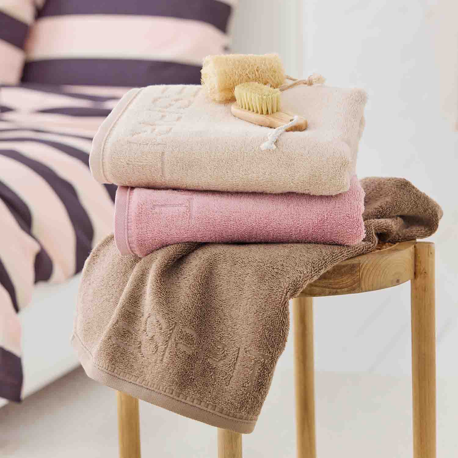 Drei Handtücher in Beige, Rosa und Braun auf Holzstuhl. Oben liegen Massagebürste und Schwamm. Im Hintergrund gestreifte Bettwäsche.