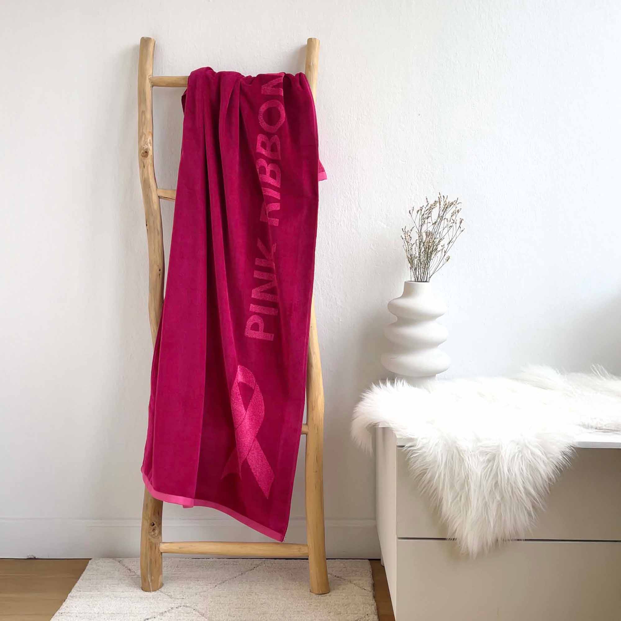 Pinkes Handtuch mit eingestickter Pink-Ribbon-Schleife hängt dekorativ an einer Holzleiter in modernem, hellen Raum – Symbol für Bewusstsein und Unterstützung.