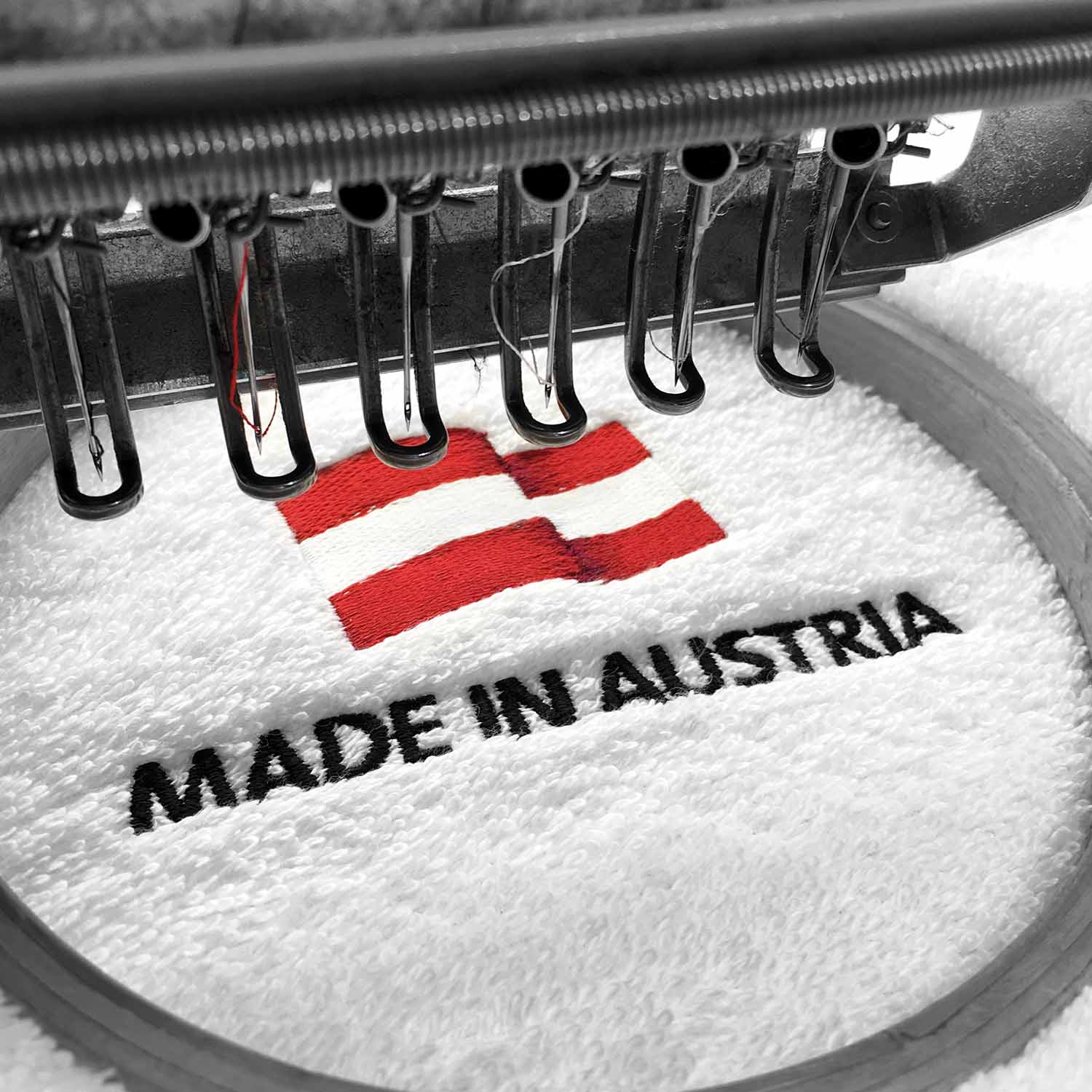 Stickmaschine stickt die österreichische Flagge mit der Unterschrift "Made in Austria" auf einem weißen Handtuch.