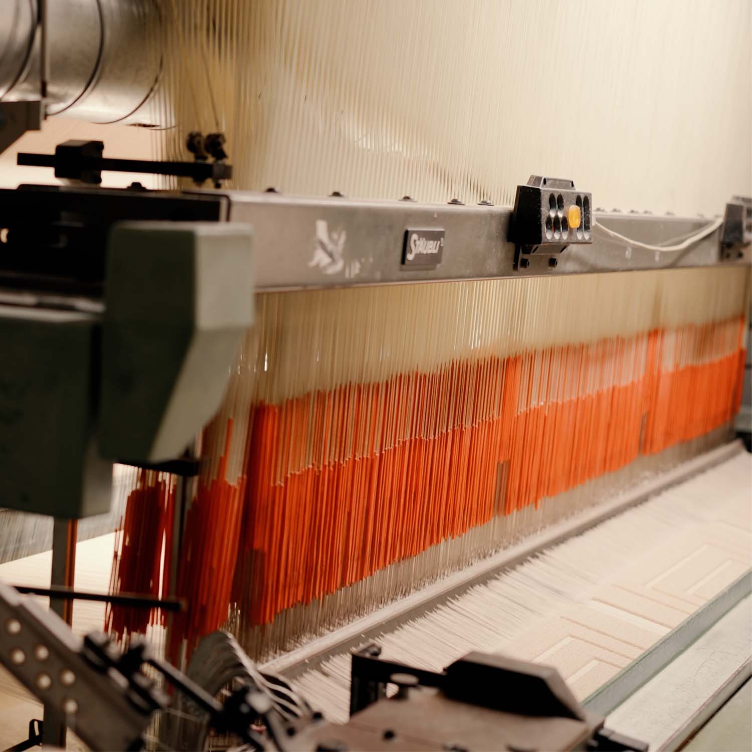 Nahaufnahme einer Webmaschine mit vertikalen Fäden, teils orange hervorgehoben, und sichtbaren mechanischen Komponenten.