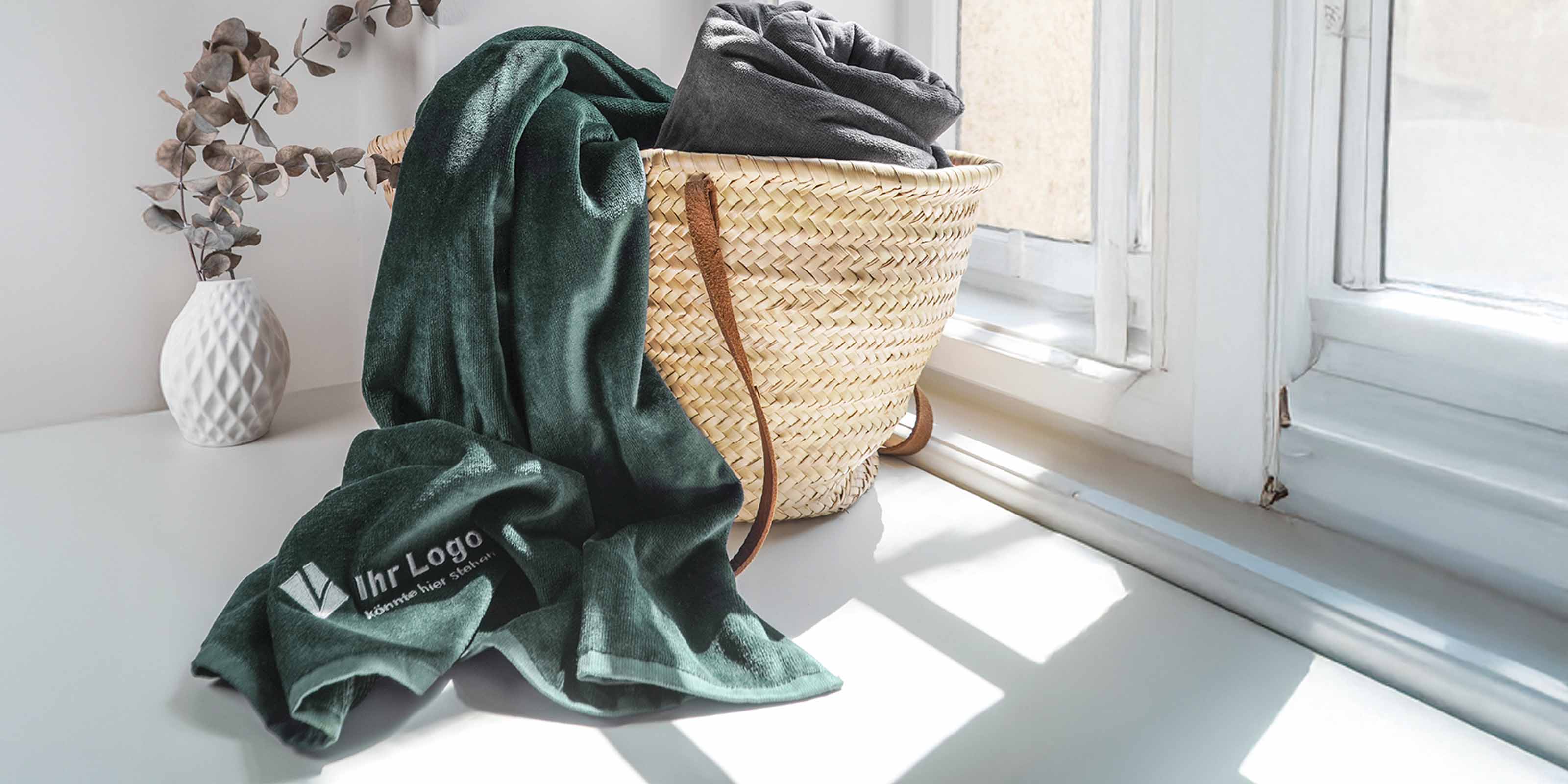 Geflochtener Korb mit grauer und grüner Decke, die grüne mit aufgesticktem Logo. Vase mit Zweigen im Hintergrund.