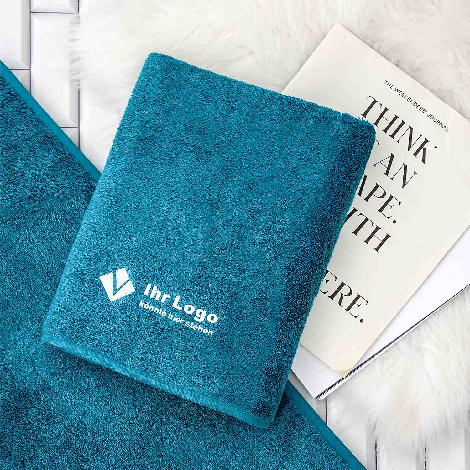 Gefaltetes blaues Handtuch mit Logo-Bestickung auf ausgebreitetem Tuch, dahinter Magazin.