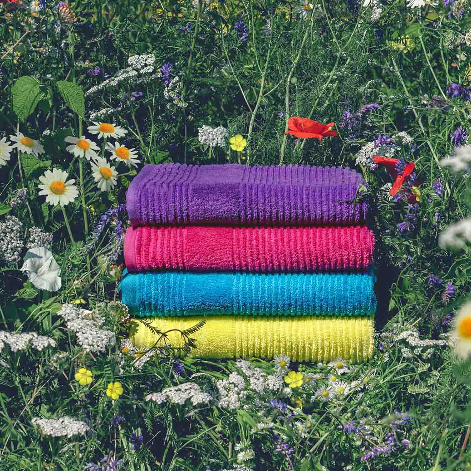 Vier farbenfrohe, gestapelte Handtücher in Lila, Pink, Blau und Gelb vor einer Wildblumenwiese. Die Szene ist mit frischen Wiesenblumen wie Gänseblümchen, Klee und anderen Wildblumen dekoriert.