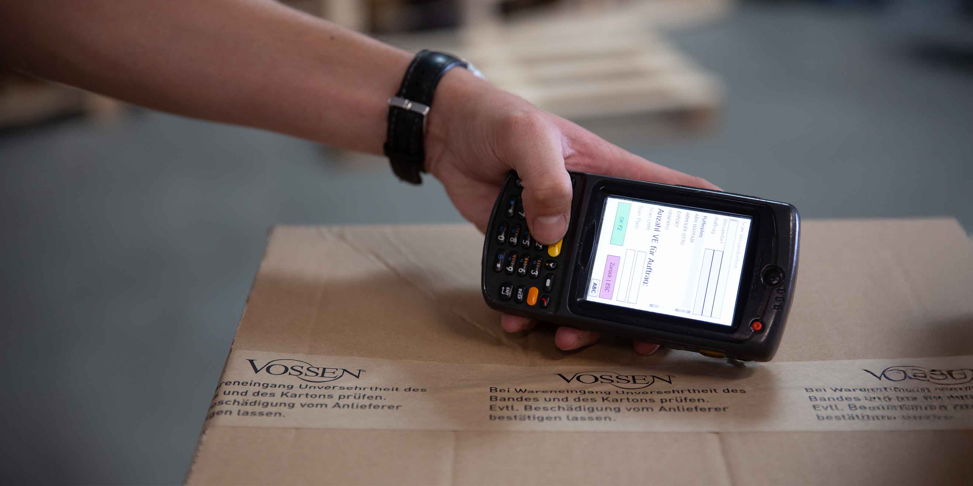 Hand mit Scannergerät über einem Karton mit VOSSEN-Logo; auf dem Gerät ist eine Eingabemaske sichtbar.