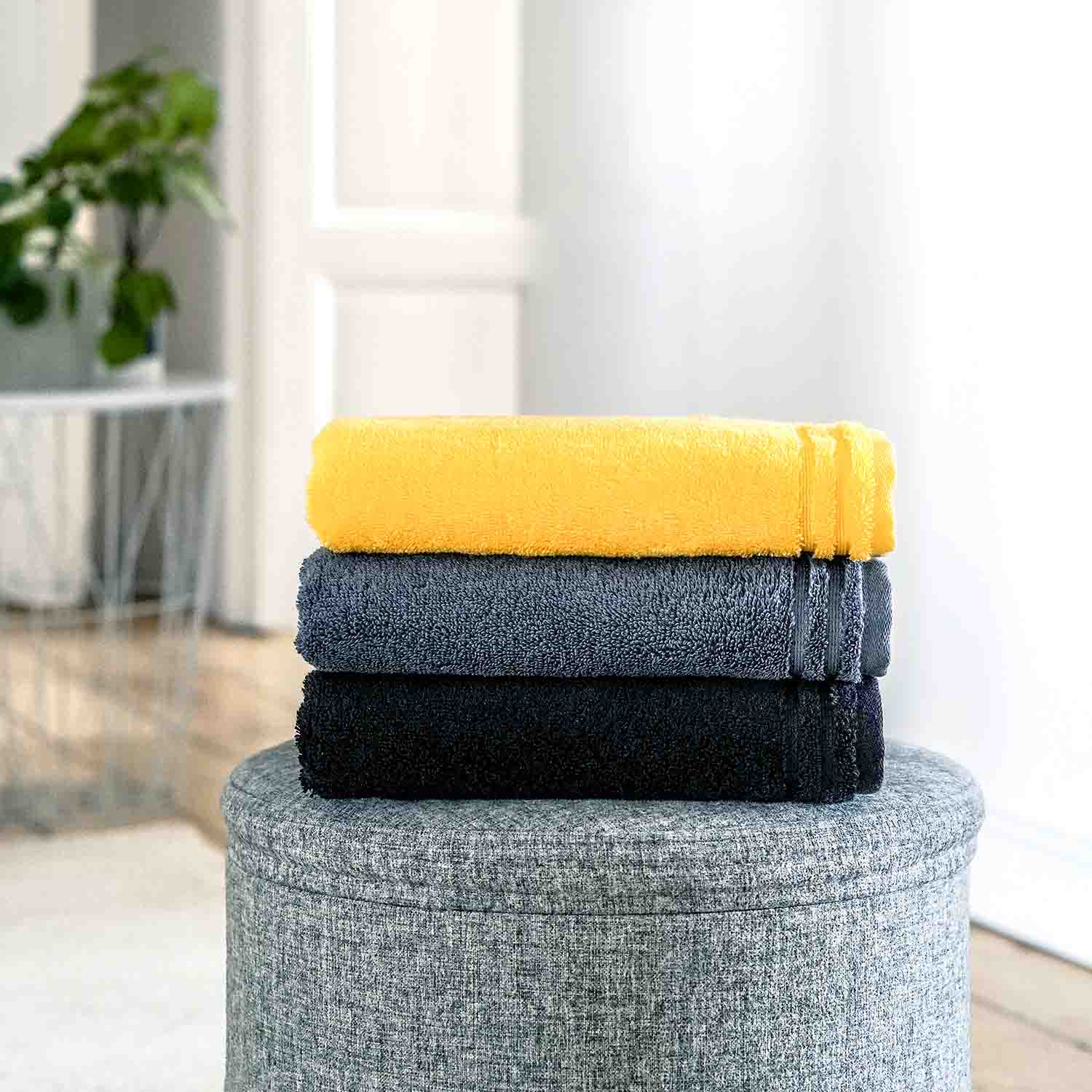 Drei gefaltete Handtücher in Gelb, Grau und Schwarz auf grauem Hocker.