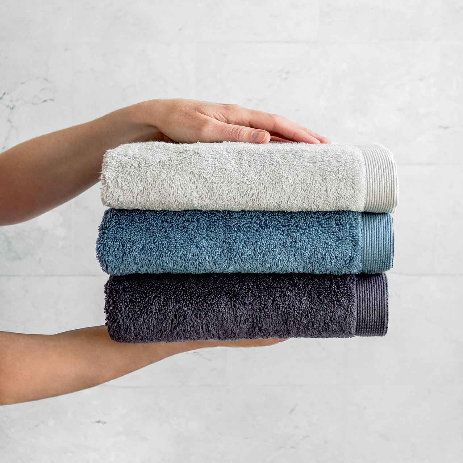 Hände halten drei gefaltete Handtücher in Grau und Blau vor hellem Hintergrund.