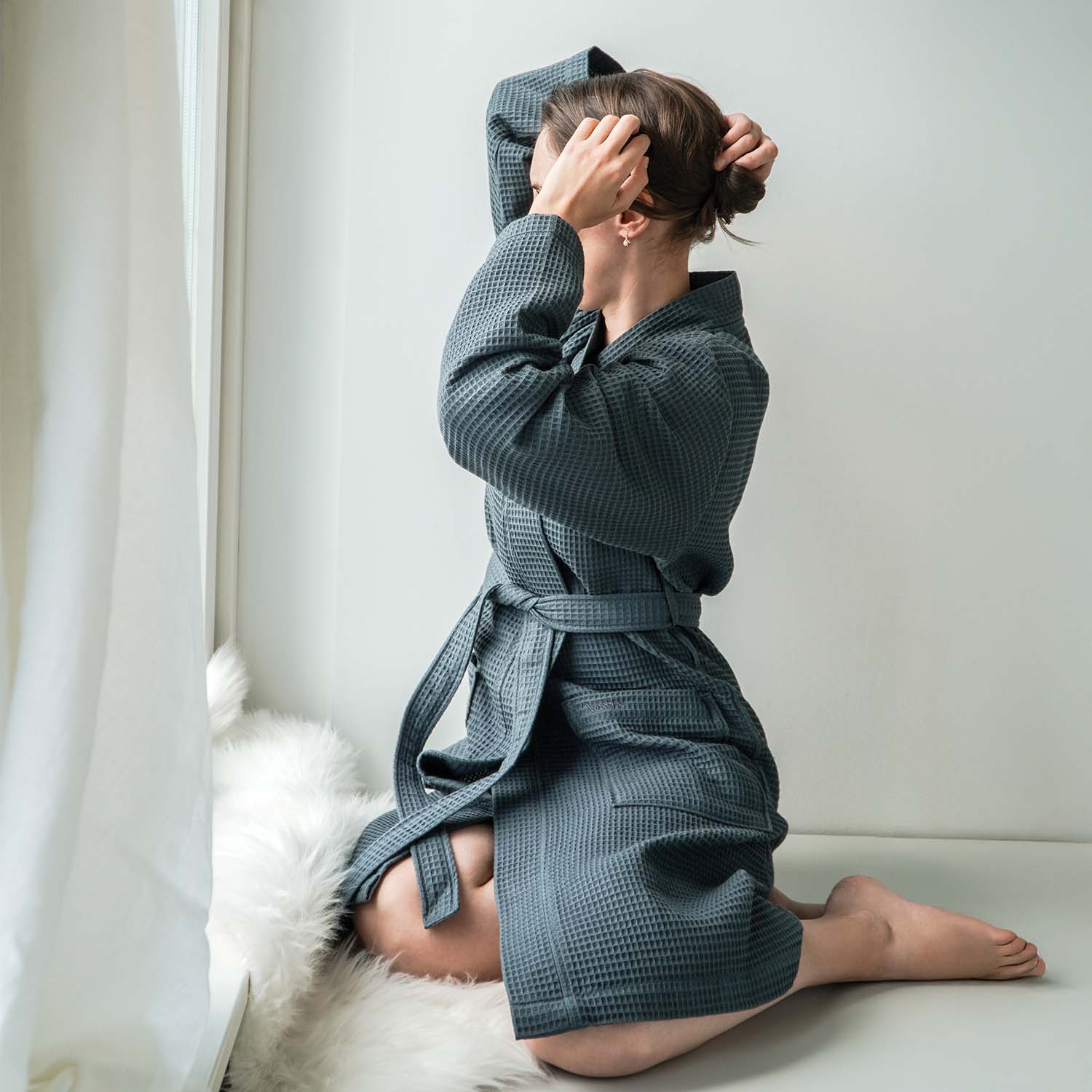 Person im grauen Bademantel kniet auf dem Boden vor weißem Hintergrund.