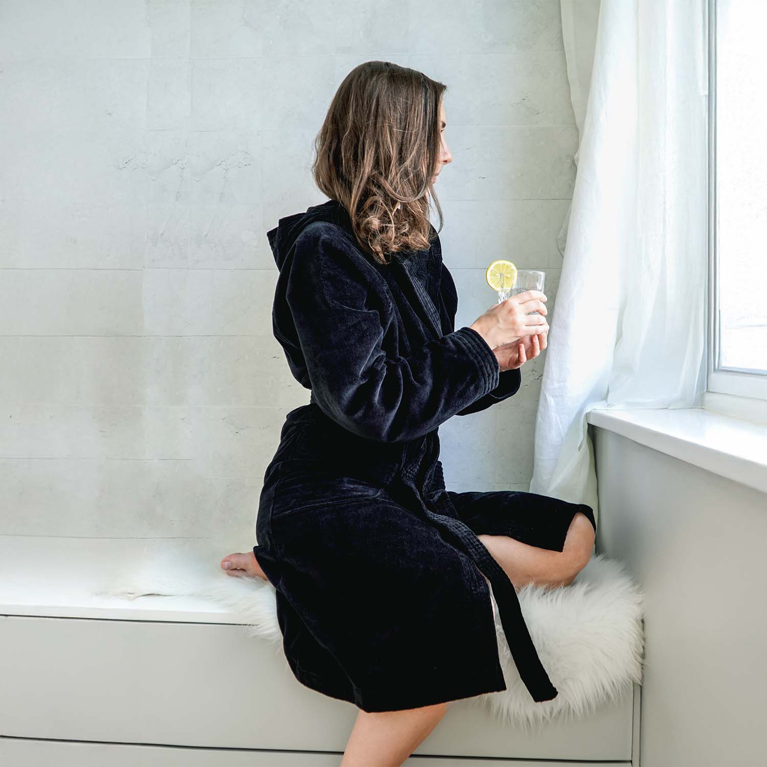Person im schwarzen Bademantel sitzt am Fenster und hält ein Glas mit einer Zitrone.