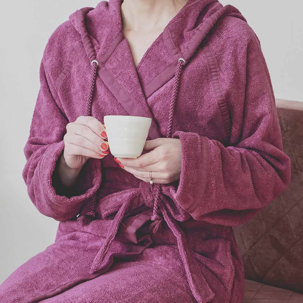 Person im lila Kapuzenbademantel hält weiße Tasse mit beiden Händen, rote Fingernägel, Ring an der linken Hand, heller Hintergrund.
