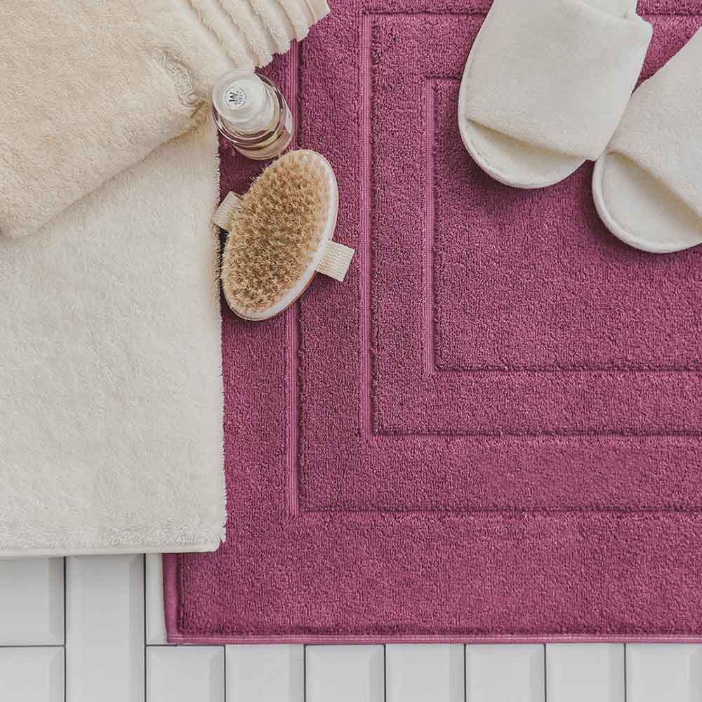 Beige Handtücher, Pflegeartikel und weiße Hausschuhe auf einem lilafarbigen Badeteppich. Alles zusammen liegt auf einem weißen Fliesenboden.