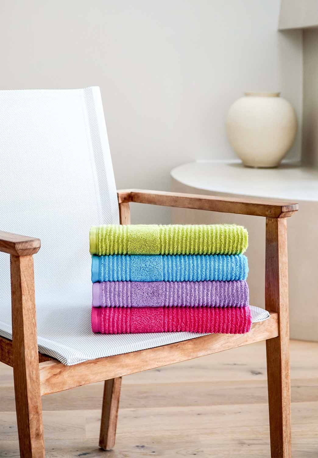 Vier gefaltete Handtücher in Grün, Blau, Lila und Rot auf weißem Stuhl mit Holzbeinen, im Hintergrund Tisch mit Vase.