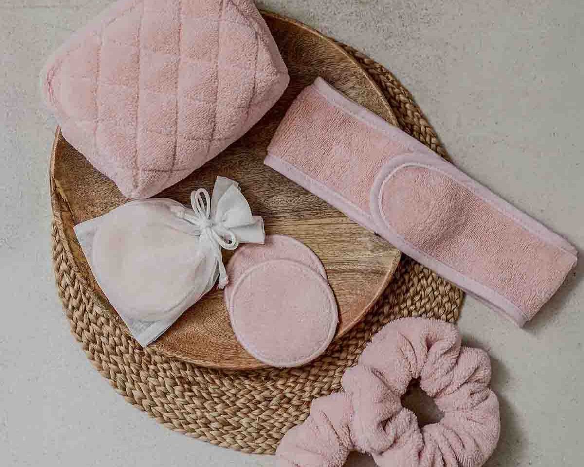 Rosa Beauty-Accessoires aus weichem Frottee – darunter ein Kosmetiktäschchen, ein Stirnband, wiederverwendbare Abschminkpads in einem Organza-Beutel und ein Haargummi – dekorativ auf einem runden Holz- und Basttablett arrangiert.