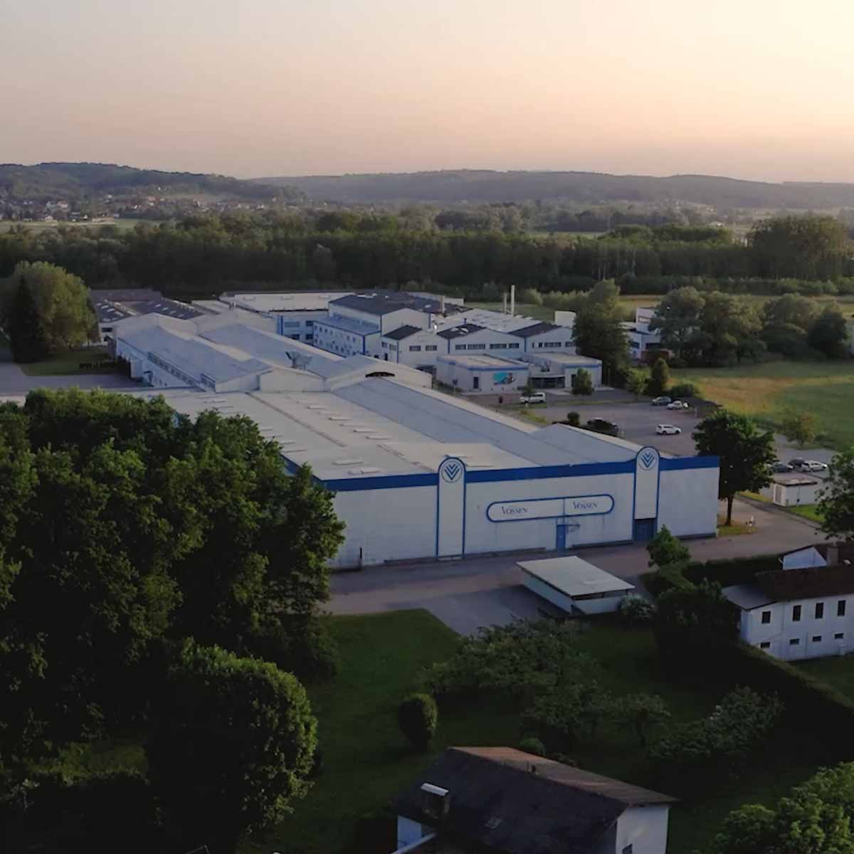 Luftaufnahme eines großen Industriekomplexes mit blauen und weißen Hallen im Grünen bei Sonnenuntergang. Auf der Fassade ist der Schriftzug „Vossen“ zu sehen. Im Hintergrund erstrecken sich Felder, Wälder und Hügel.