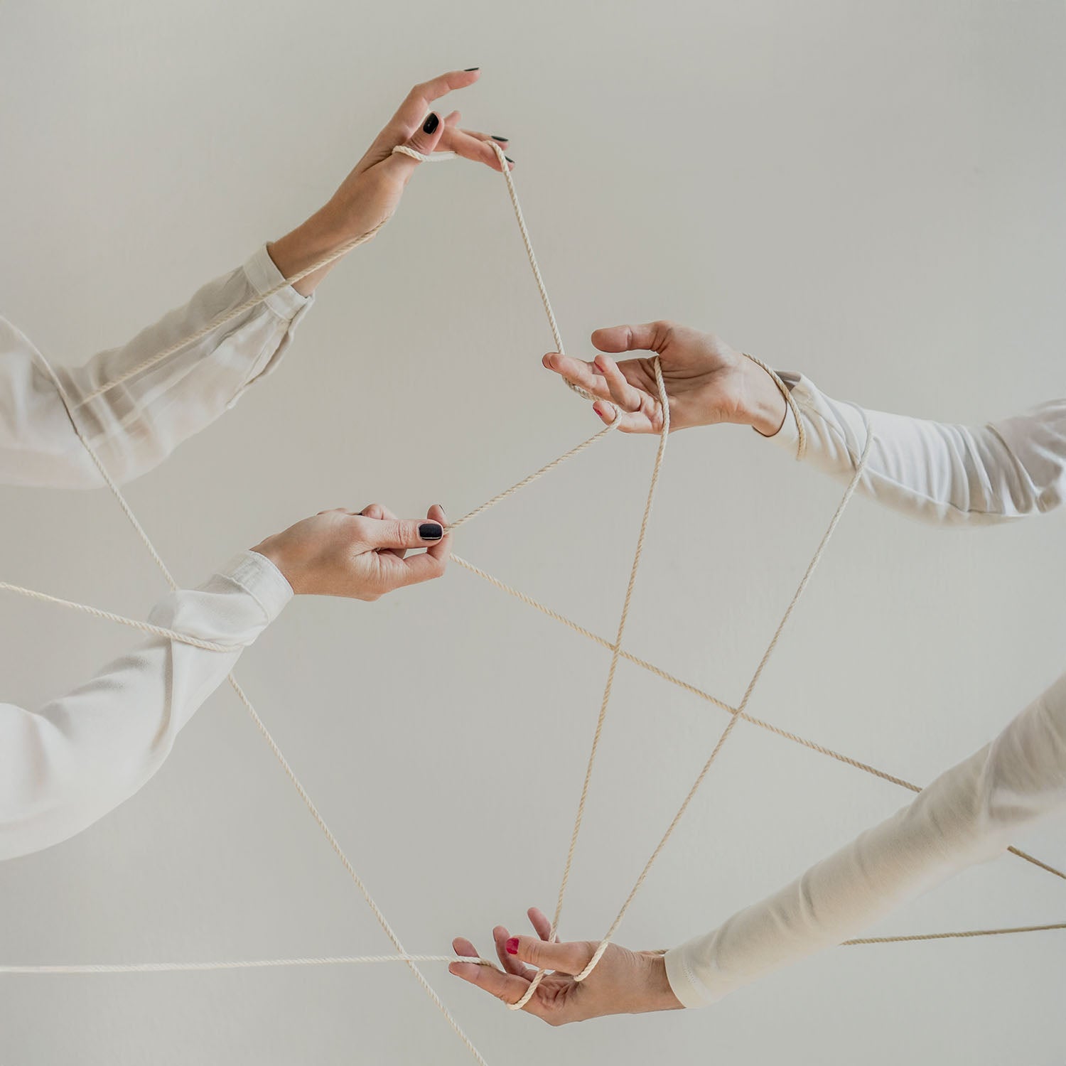 Vier Hände spielen mit weißen Fäden ein Fadenspiel vor hellem Hintergrund.