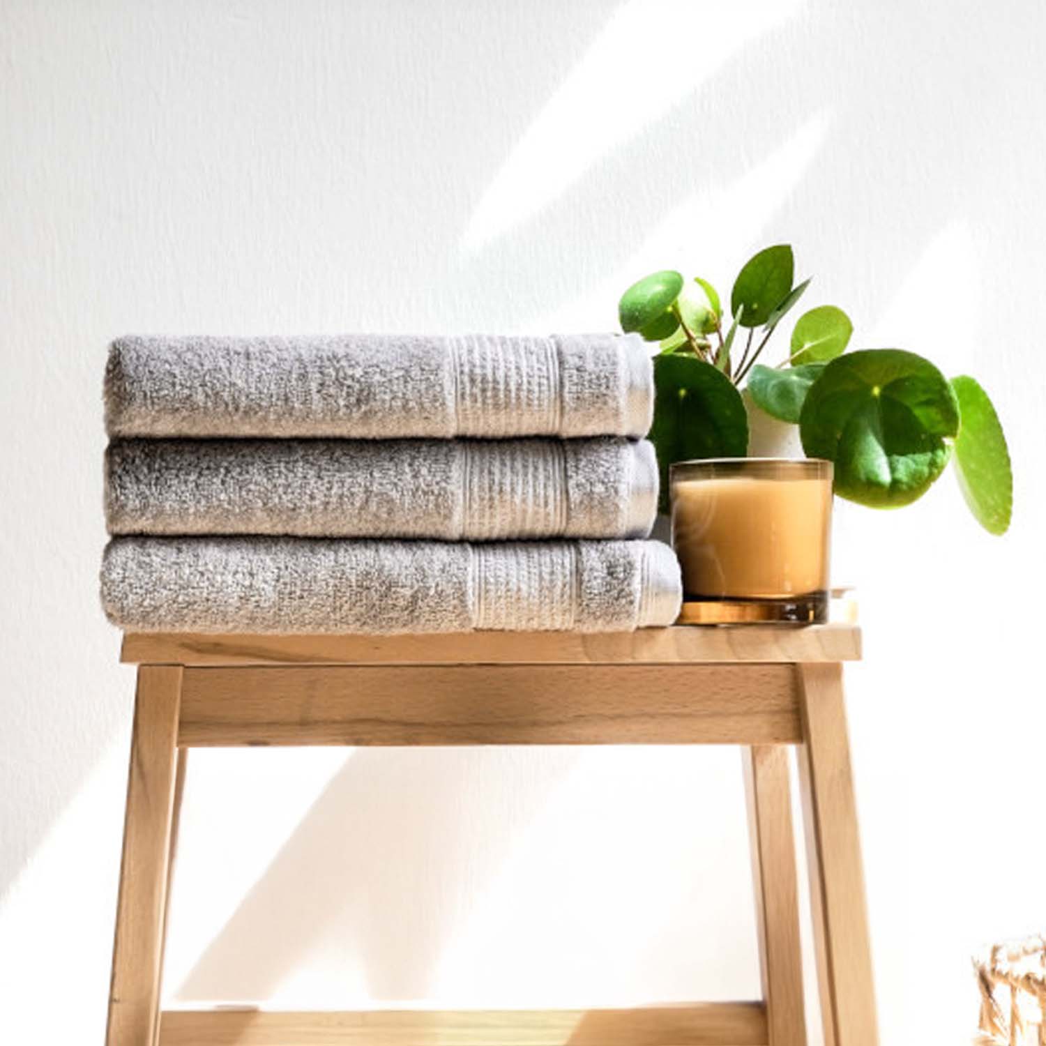 Drei graue, gefaltete Handtücher liegen ordentlich auf einem hellen Holzhocker. Daneben stehen eine kleine grüne Pflanze und eine beigefarbene Kerze im Sonnenlicht.