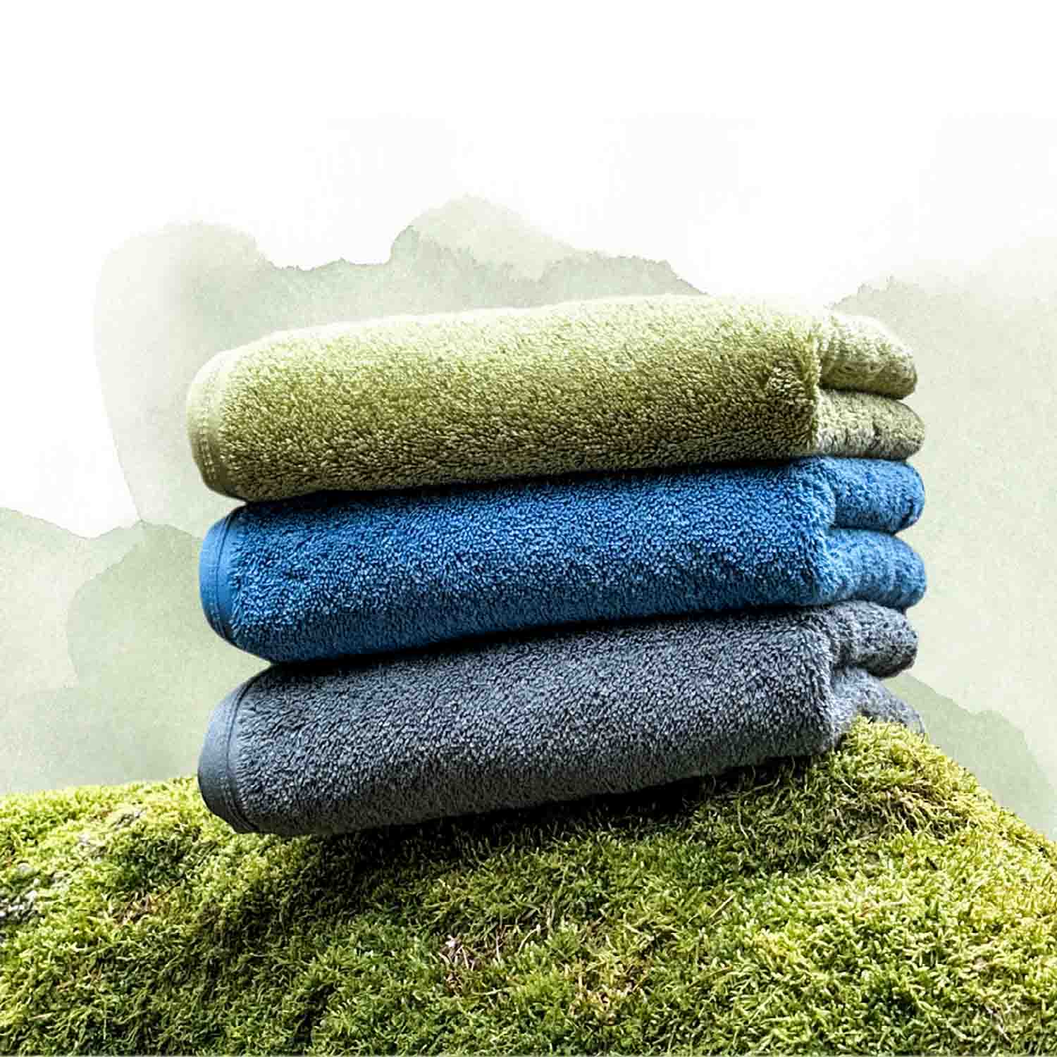 Drei farbige Handtücher in Grün, Blau und Grau liegen gestapelt auf einer moosbedeckten Fläche vor einem hellen, natürlichen Hintergrund.