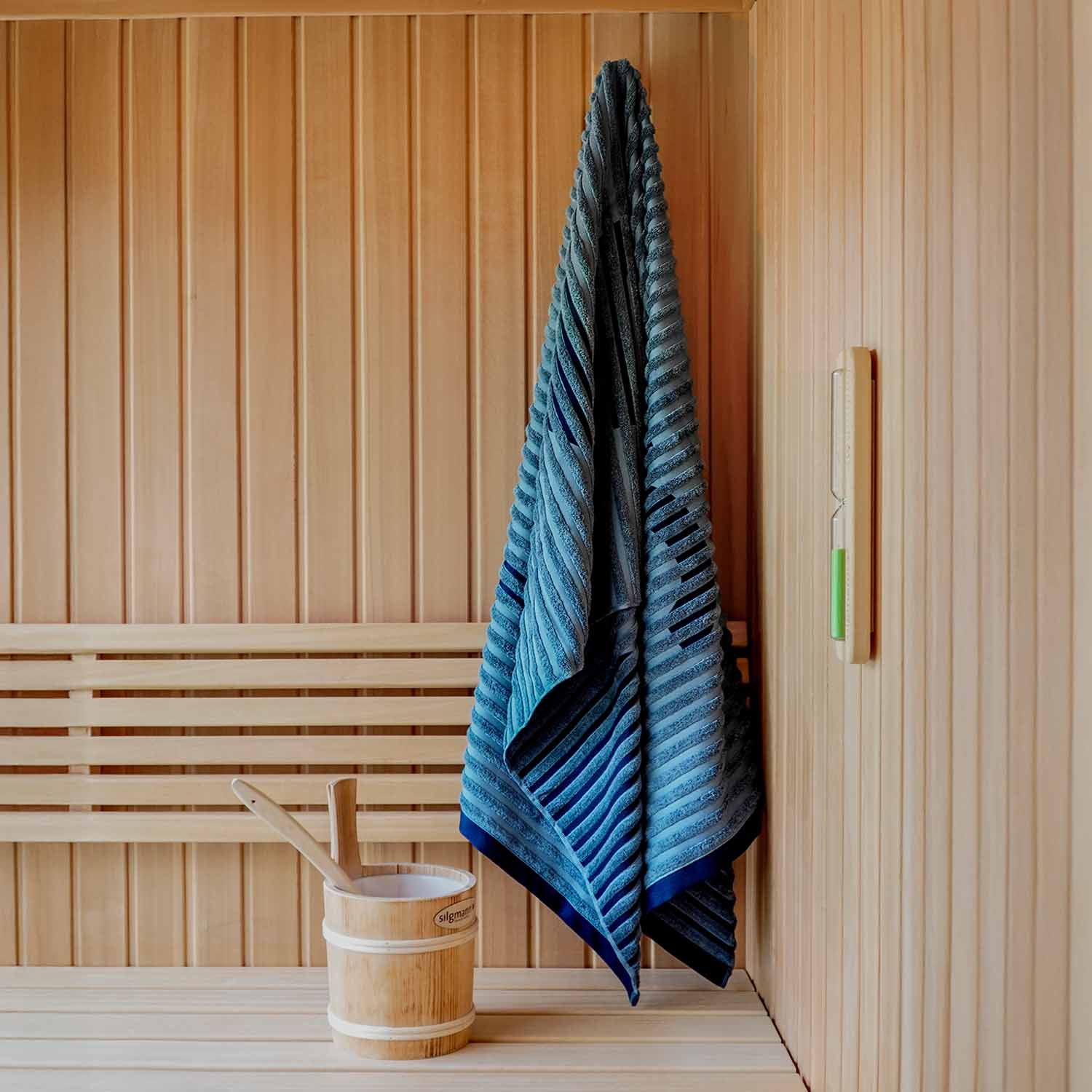 Innenraum einer Sauna mit Holzbank. Auf der Bank stehen ein Holzeimer mit zwei Schöpfkellen und ein blaues Handtuch hängt an der Wand.