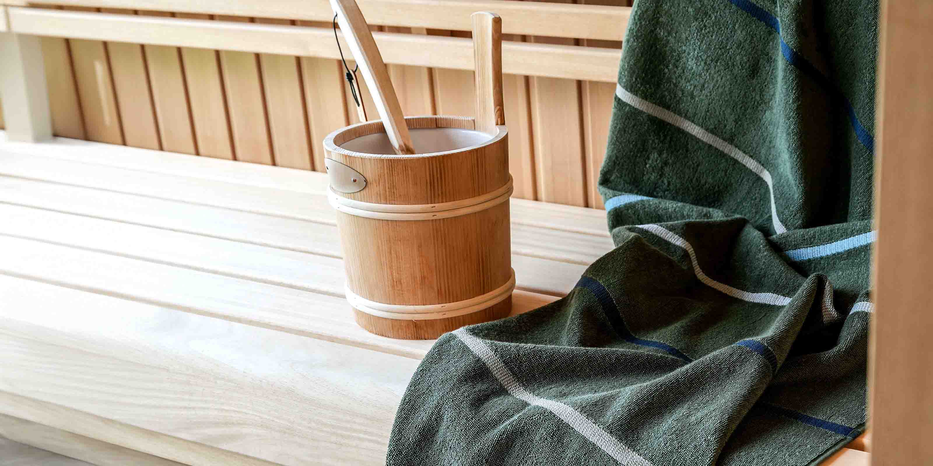 Grünes Handtuch mit Streifen auf Holzbank neben Saunaeimer mit zwei Schöpfkellen.