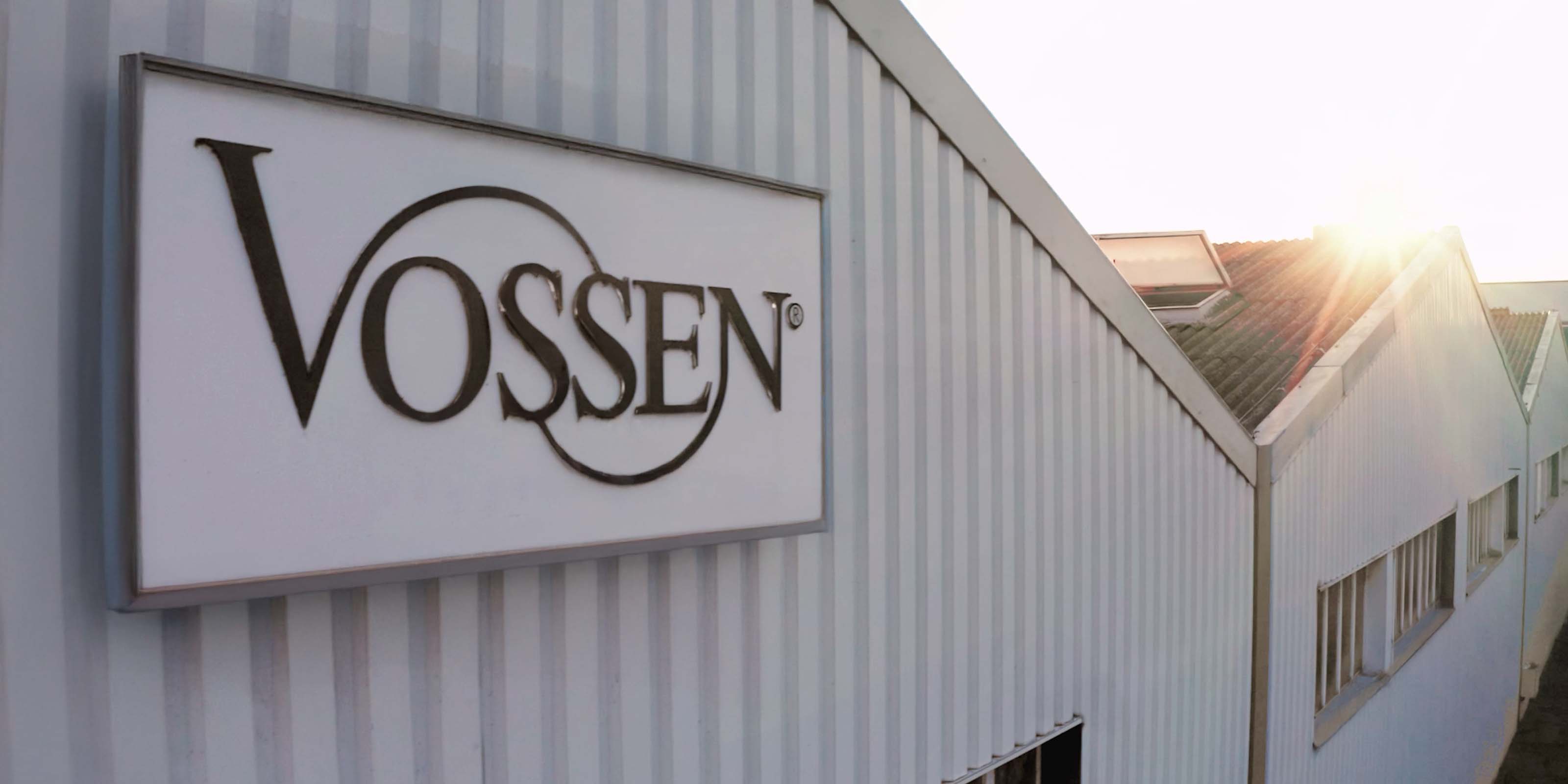 Nahaufnahme eines Gebäudes mit dem großen Firmenlogo „VOSSEN“ an der Fassade; im Hintergrund scheint die Sonne über das Dach der Industriehalle.