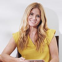 Person mit langen, welligen blonden Haaren und gelbem ärmellosem Oberteil hält ein Objekt, vermutlich Tablet oder Buch. Hintergrund hell und schlicht.