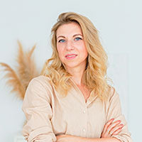 Person mit langen, welligen blonden Haaren und hellem Shirt vor Hintergrund mit getrockneten Pflanzen.