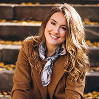 Person mit langen, welligen Haaren sitzt auf Außentreppe mit Herbstlaub. Trägt braunen Mantel und Schal.