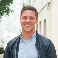 Person mit hellblauem Hemd und dunkler Jacke steht draußen in städtischer Umgebung. Im Hintergrund Gebäude und Bäume.