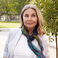 Person mit weißem Hemd, hellem Blazer und buntem Schal steht draußen vor grüner Kulisse.