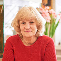 Person mit blondem Haar und rotem Pullover vor unscharfem Hintergrund mit Blumen.