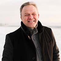 Person mit dunklem Mantel über einem Strickpullover steht draußen vor unscharfem Hintergrund.