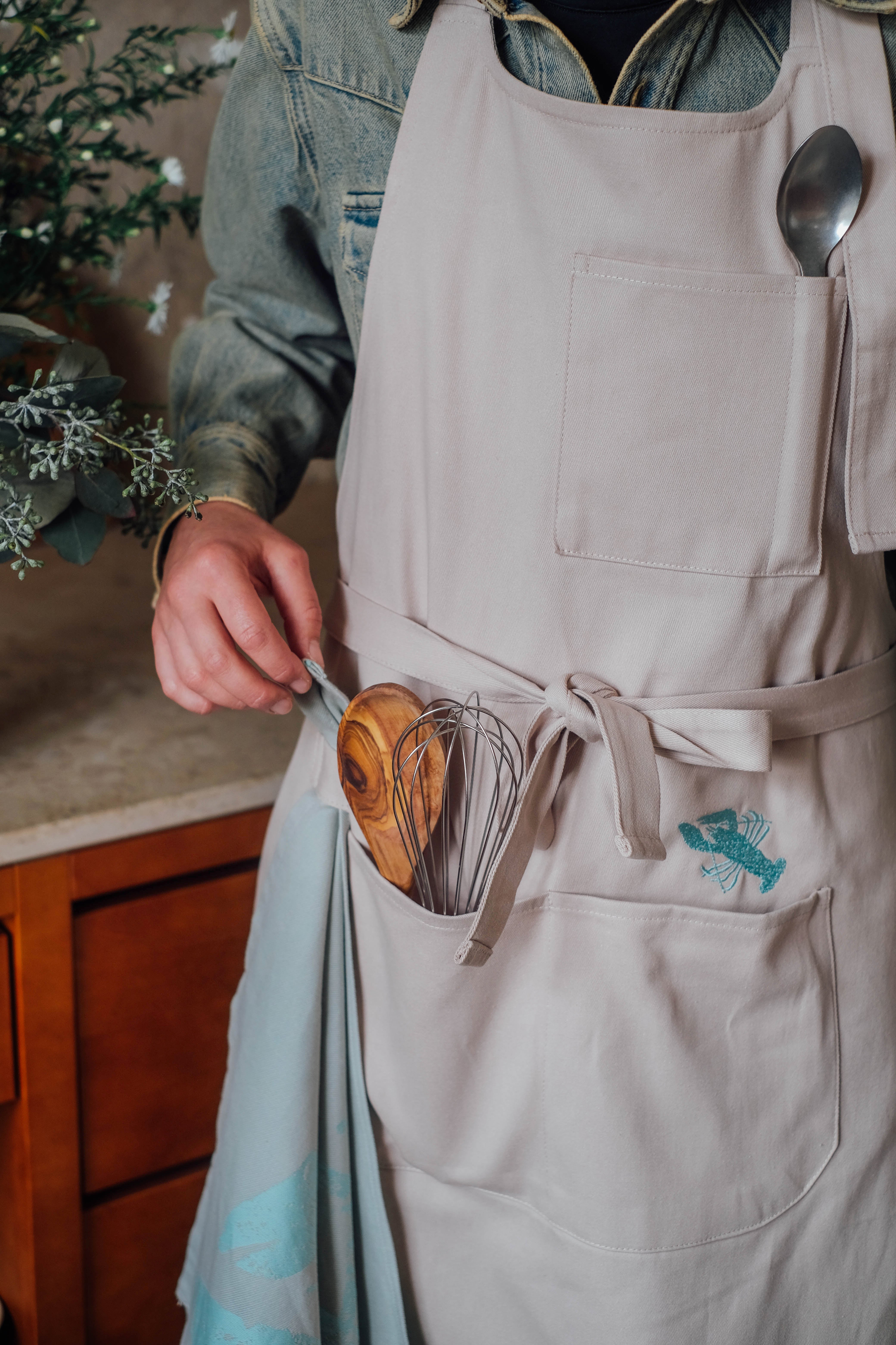 Person mit heller Küchenschürze und Jeansjacke in einer Küche. In den Taschen der Schürze befinden sich Kochlöffel, Schneebesen und Besteck. Seitlich hängt ein Küchenhandtuch in Mint mit abstraktem Muster.