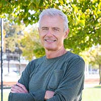 Person mit grauem Haar steht draußen mit verschränkten Armen. Im Hintergrund grüne Bäume und Gebäude.