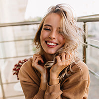 Person mit langen Haaren und braunem Sweatshirt sitzt auf einem Balkon oder einer Terrasse. Im Hintergrund Geländer und Pflanzen.