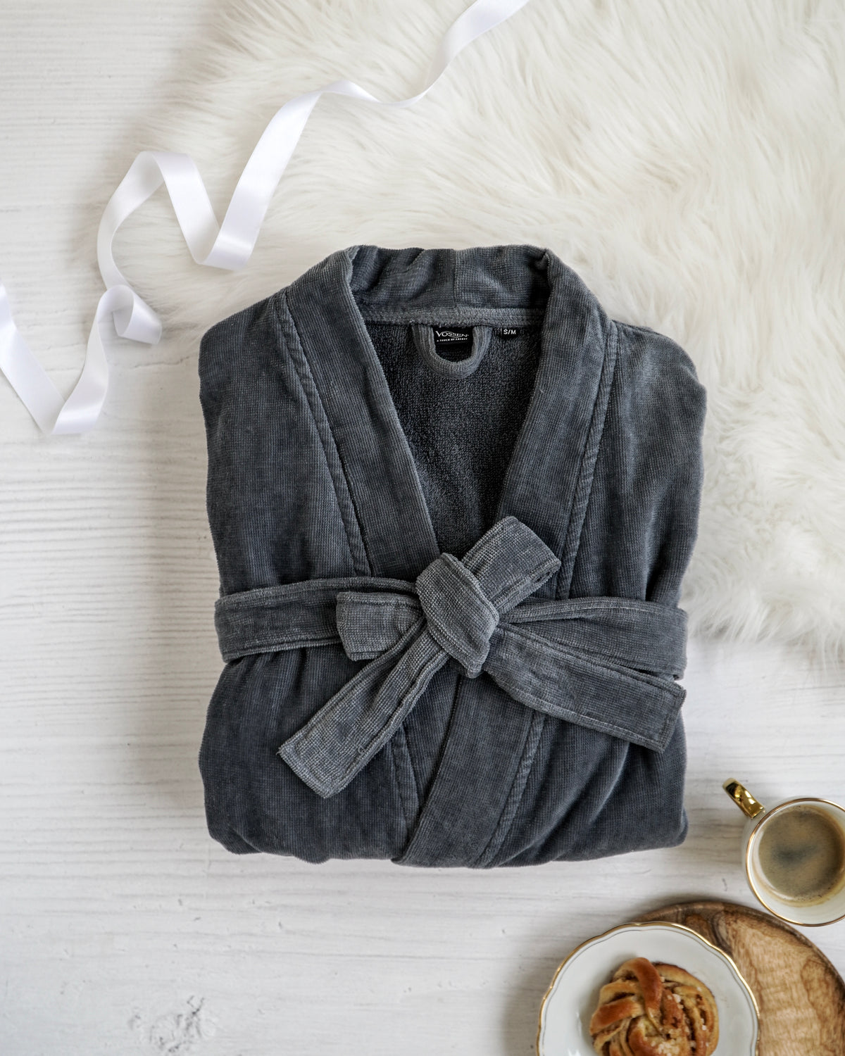 Ein elegant gefalteter, dunkelgrauer VOSSEN Bademantel mit Bindegürtel liegt auf einer hellen Oberfläche neben einem weißen Fell, dekorativem Geschenkband und einer Tasse Kaffee. Die stilvolle Anordnung vermittelt Gemütlichkeit und luxuriösen Wellnesskomfort.
