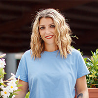 Person mit schulterlangem, welligem blondem Haar und hellblauem T-Shirt steht draußen vor grüner Kulisse mit Blumen.