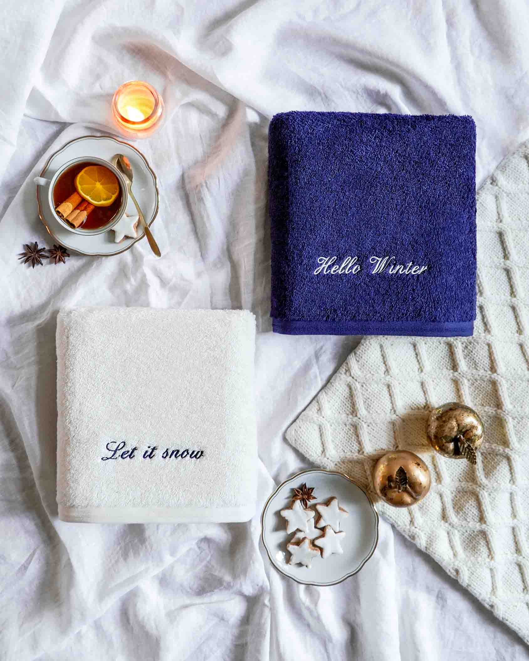 Winterliche VOSSEN Handtücher in Weiß und Blau mit Stickerei ‚Let it snow‘ und ‚Hello Winter‘, liebevoll dekoriert mit Tee, Zimtsternen und Weihnachtskugeln – perfekte Geschenkidee für die kalte Jahreszeit.