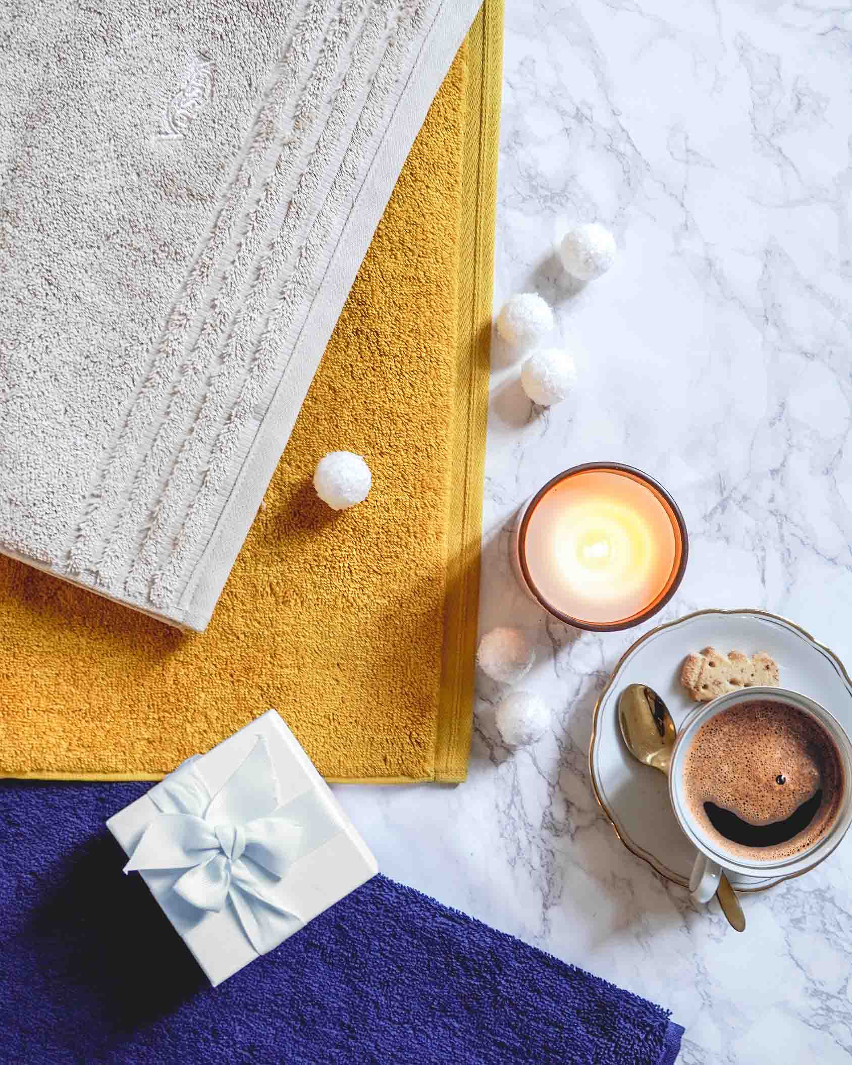 Farbenfrohe VOSSEN Handtücher in Beige, Senfgelb und Dunkelblau, dekorativ arrangiert mit Geschenkbox, Kerze und Kaffee – stilvolle Geschenkidee für Wellnessliebhaber.