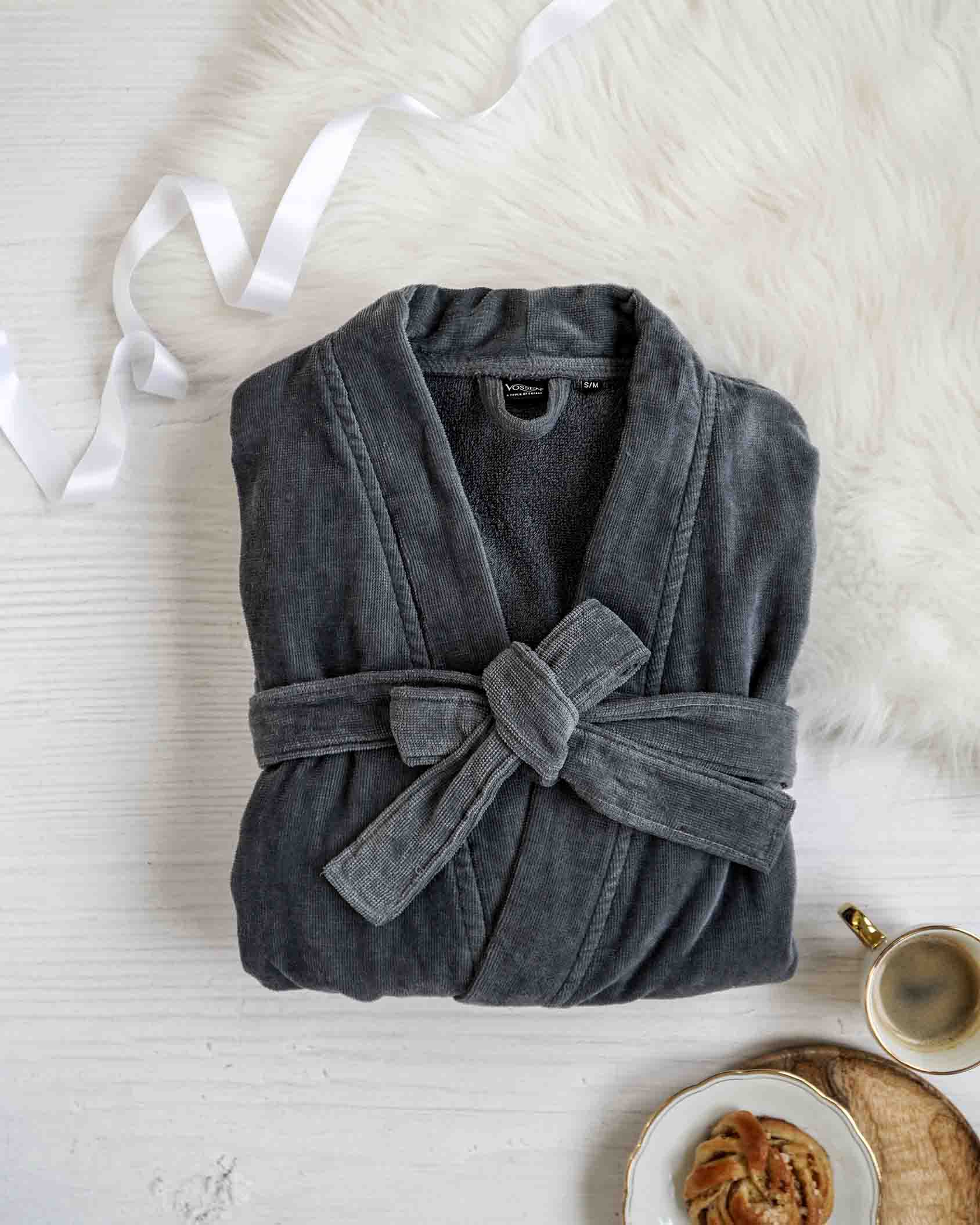 Gefalteter grauer VOSSEN Bademantel aus weichem Baumwollmaterial, liebevoll arrangiert auf weißer Fläche mit Schleife, Kaffee und Gebäck – perfektes Wellness-Geschenk für Zuhause.