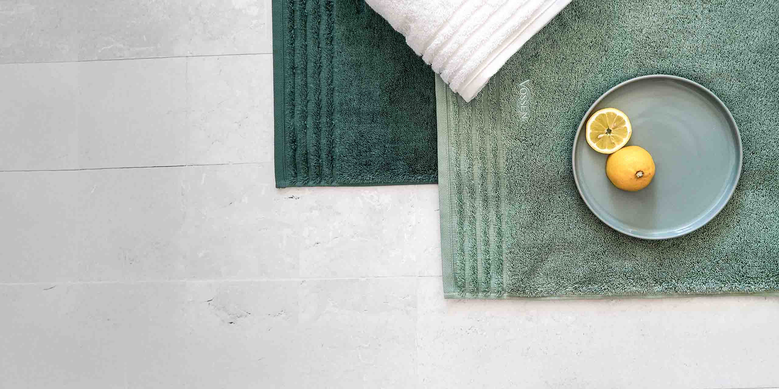 Zwei grüne Handtücher auf hellem Boden, darauf ein weißes Handtuch und ein Teller mit Zitronen.