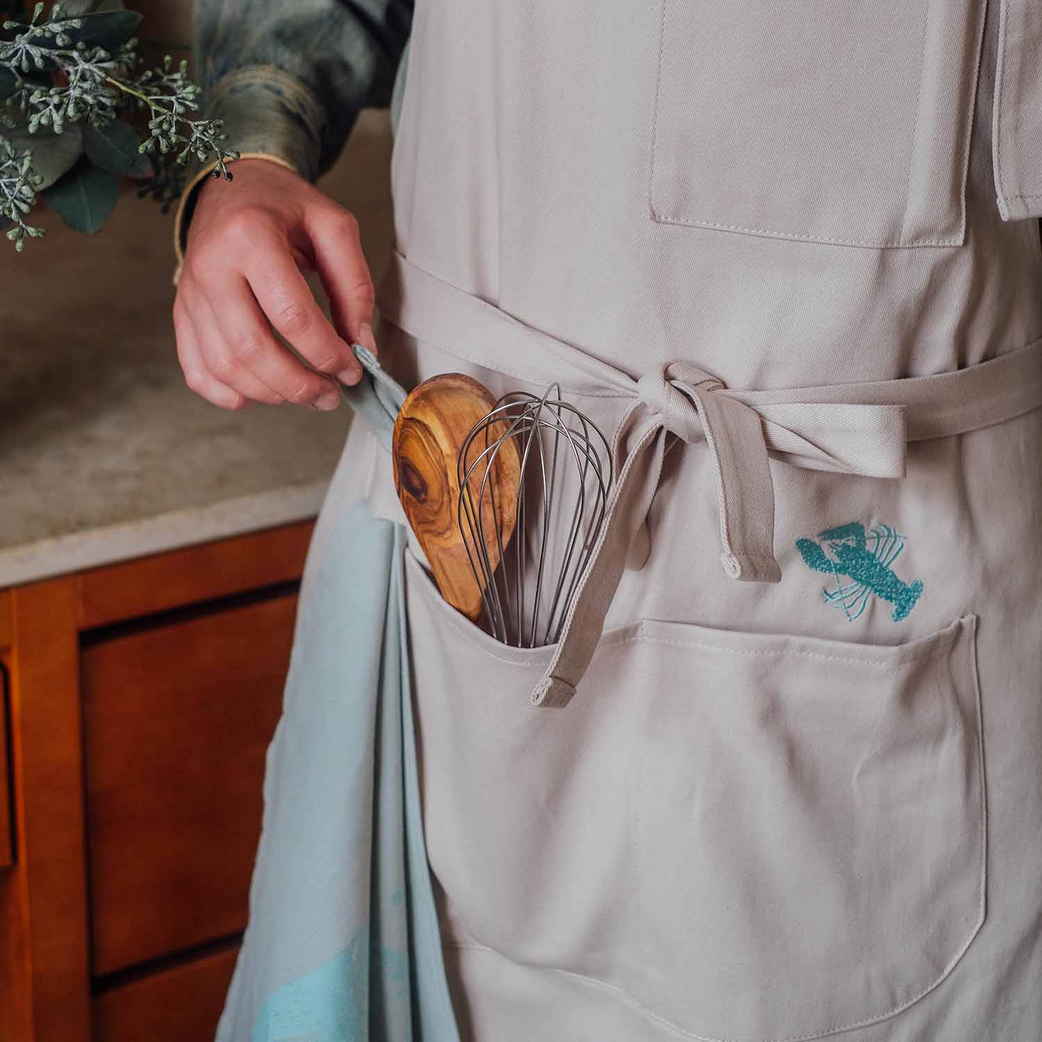 Beige Küchen-Schürze mit aufgesticktem Hummer-Motiv und Kochutensilien in der Tasche, Person hält ein Geschirrtuch