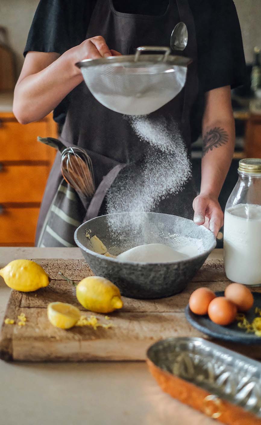 Person siebt Mehl in eine Rührschüssel beim Backen von Kuchen, mit frischen Zitronen, Eiern und Milch auf der Küchenarbeitsplatte