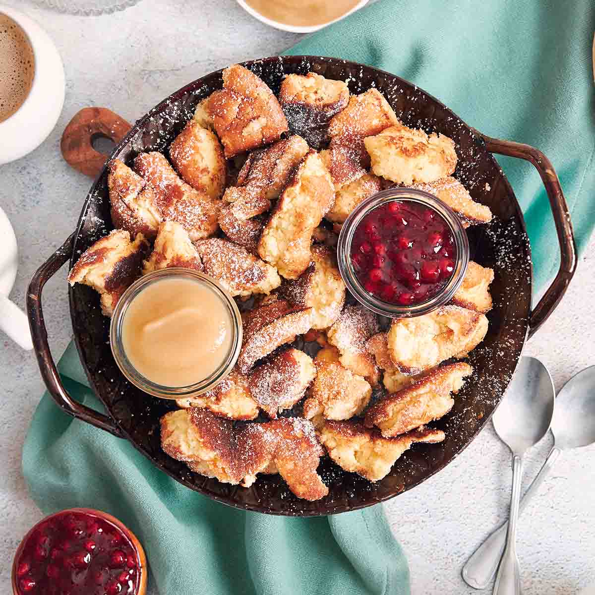 Pfanne mit Kaiserschmarrn, bestäubt mit Puderzucker und serviert mit Vanillesauce und roter Beerenkompott-Dip, daneben Kaffee und Löffel