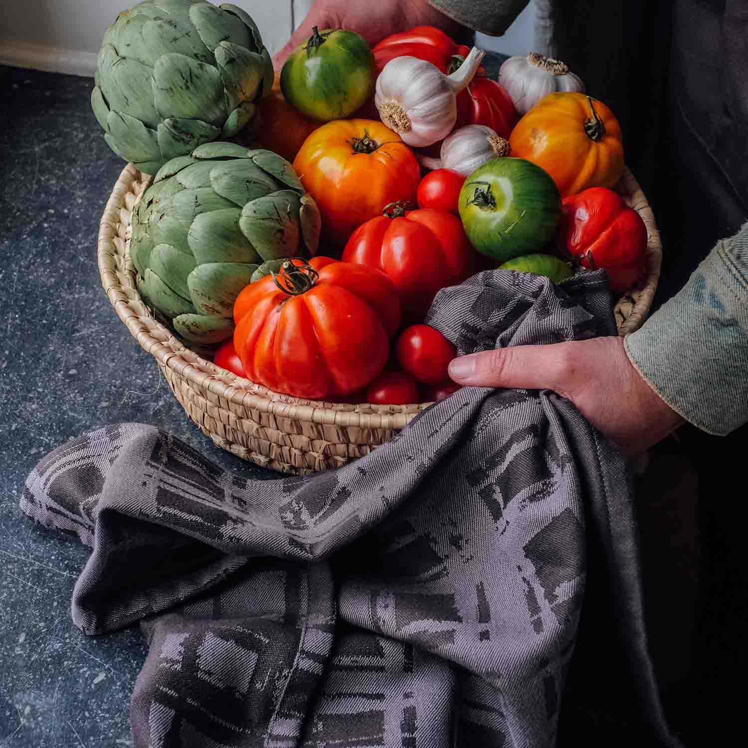 Geflochtener Korb mit frischem Gemüse wie Artischocken, Tomaten und Knoblauch auf Küchenarbeitsplatte – nachhaltiges Kochen mit hochwertigem Küchentuch aus Baumwolle