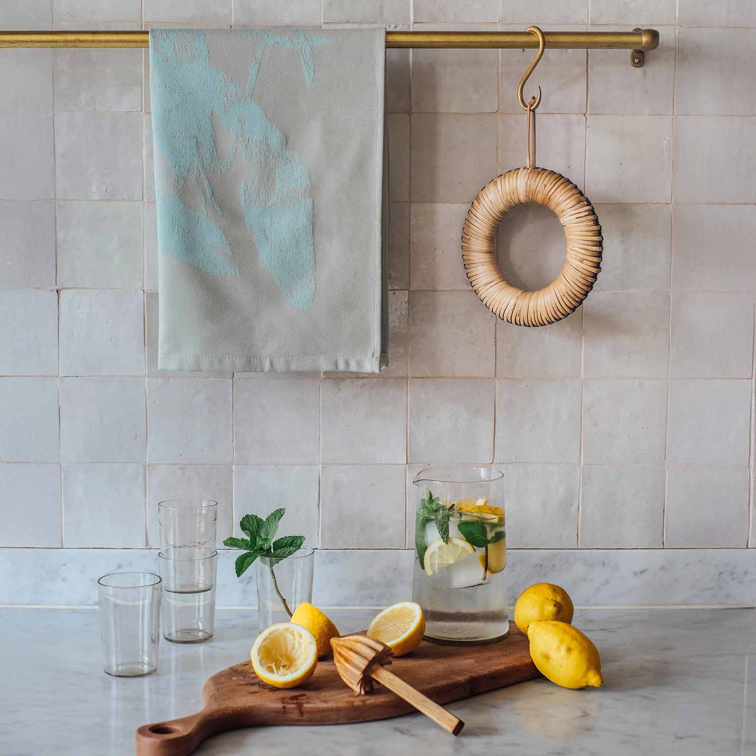 Modernes Geschirrtuch in Mintgrün an goldener Küchenstange, darunter Zitronen und Glas mit Zitronenwasser auf Arbeitsplatte