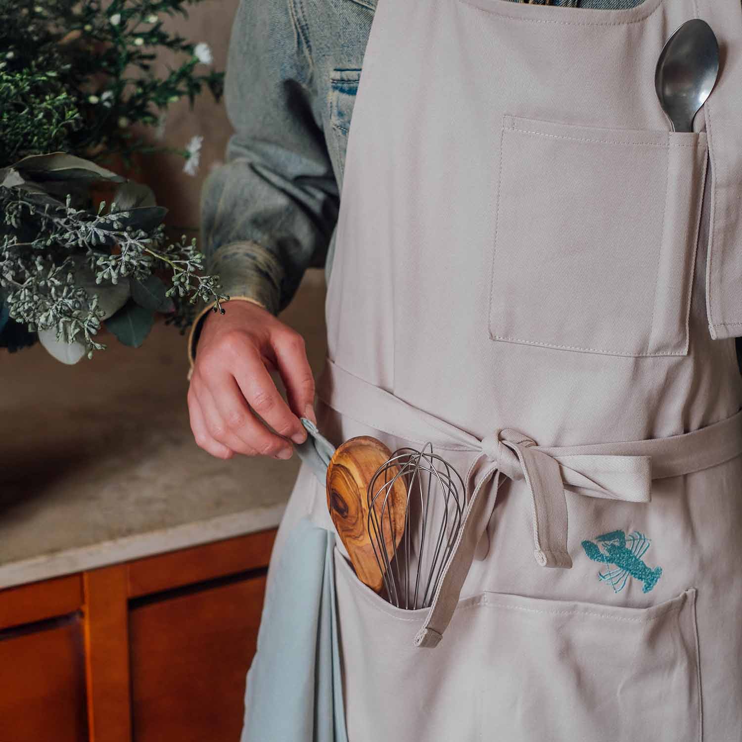 Beige Küchen-Schürze mit aufgesticktem Hummer-Motiv und Kochutensilien in der Tasche, Person hält ein Geschirrtuch