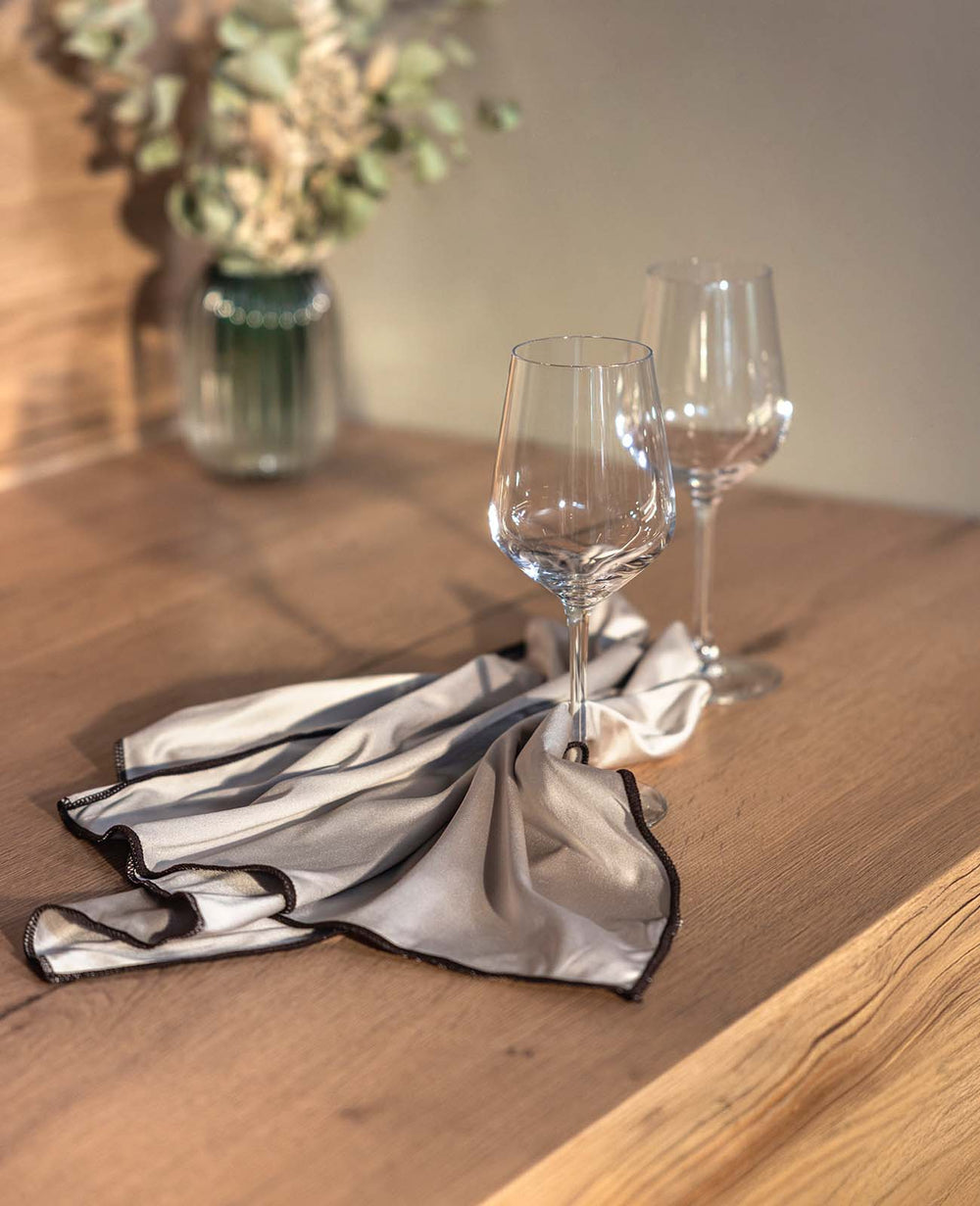 Zwei elegante Weingläser auf einem Holztisch, dekorativ arrangiert auf einem hellbeigen Tuch mit dunkler Einfassung. Warmes Sonnenlicht sorgt für eine gemütliche Atmosphäre.